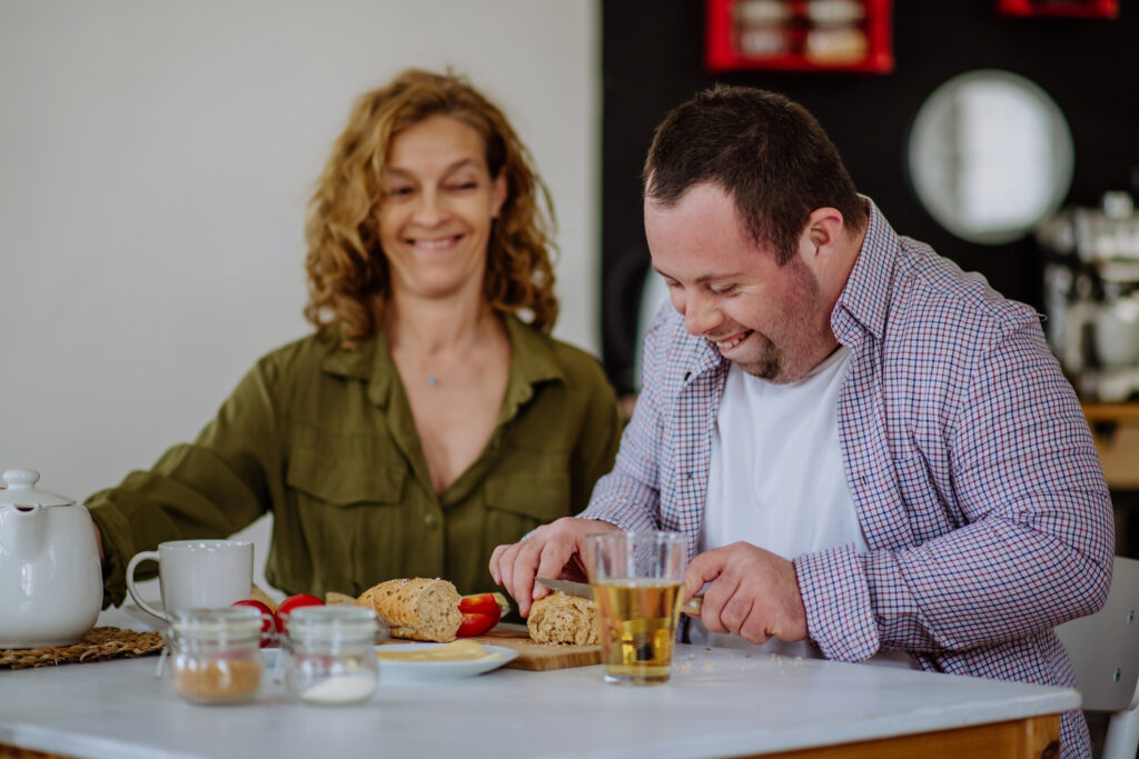 A man with Downs Syndrome works at a kitchen bench, smiling, while a support worker watches on.