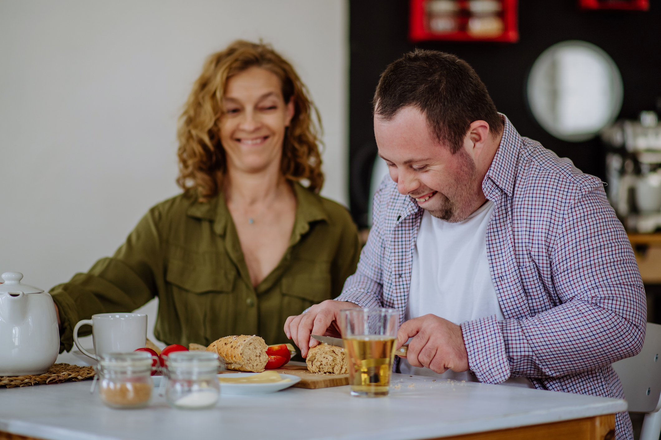 A man with Downs Syndrome works at a kitchen bench, smiling, while a support worker watches on.